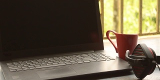 带麦克风的耳机放在靠近笔记本电脑的地方，在早晨的阳光照射下。红杯咖啡在远处。呆在家里，在家工作，远程工作，业务连续性背景。
