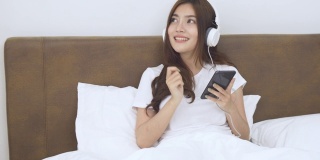 亚洲快乐的年轻女子戴耳机使用智能手机躺在床上在家放松移动的手，有乐趣。生活方式