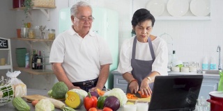 一对亚洲老年夫妇在家做饭时使用笔记本电脑。70年代老年男女关系退休生活方式概念