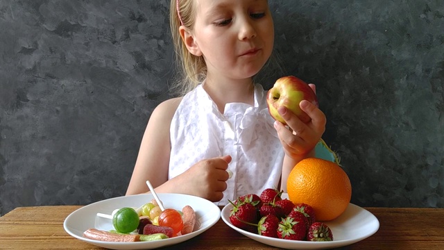 小女孩在家里的桌子上放着新鲜的水果和糖果。孩子们认为该选择健康的还是有害的食物。高清