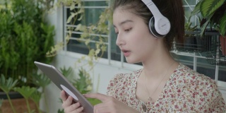 2019冠状病毒病(COVID-19)疫情发生后，一名年轻的亚洲女性在被隔离期间，手持平板电子设备在起居室里通过耳机听音乐，在温室的小花园中放松，室内gar的概念