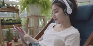 2019冠状病毒病(COVID-19)疫情发生后，一名年轻的亚洲女性在被隔离期间，手持平板电子设备在起居室里通过耳机听音乐，在温室的小花园中放松，室内gar的概念