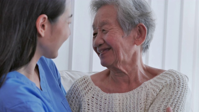 亚洲老年妇女听亚洲年轻女护士医生在咨询和拥抱她的年长女性病人在家里。在家里照顾者