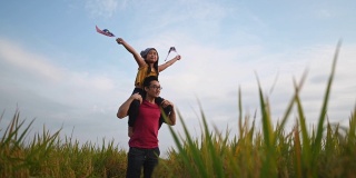 马来西亚独立日，一个亚洲华人小女孩在操场上举着马来西亚国旗，由她的父亲扛在她父亲的肩膀上，享受着清晨的阳光，感到自豪和快乐