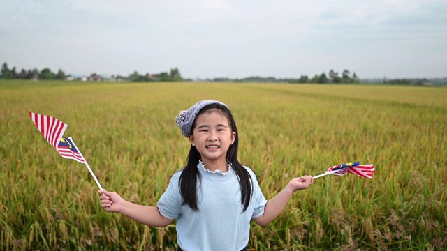 马来西亚独立日当天，一名亚洲华裔女孩在操场上挥舞着马来西亚国旗，享受着清晨的阳光，感到自豪和快乐