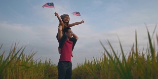 马来西亚独立日，一个亚洲华人小女孩在操场上举着马来西亚国旗，由她的父亲扛在她父亲的肩膀上，享受着清晨的阳光，感到自豪和快乐