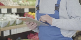 穿着制服的杂货店男员工在货架上走着，用着平板电脑。成年人在零售店检查水果和蔬菜的清单。商业、消费、生活方式。S-log 2。