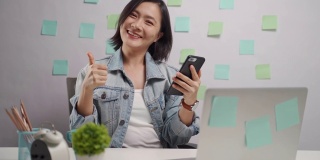亚洲妇女高兴地看智能手机阅读新闻显示拇指在家庭办公室。亲密。居家办公