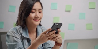 亚洲女人快乐微笑看智能手机和阅读新闻在家办公室。