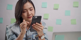亚洲女人快乐微笑看智能手机和阅读新闻在家办公室。