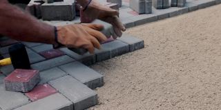 一位戴着手套的工匠正在用气泡水平仪和橡胶锤铺设混凝土砖。瓷砖铺设的声音。砖人行道铺设的专业摊铺机。在院子里铺上灰色的混凝土铺路板