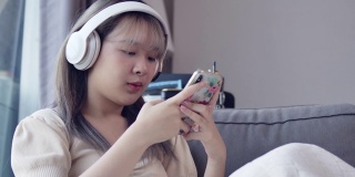 一个美丽的亚洲少女听音乐耳机和智能手机在她的客厅在公寓房间。