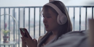 一个美丽的亚洲少女听音乐耳机和智能手机在她的客厅在公寓房间。
