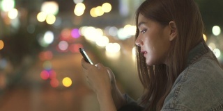 年轻美丽的亚洲女性微笑着使用智能手机上网社交媒体或网上购物，看着城市的夜光。漂亮女孩喜欢城市夜生活。