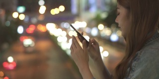 微笑的年轻美丽的亚洲女人用智能手机上网社交媒体或网上购物，并拍下城市的路灯。快乐的漂亮女孩喜欢城市夜生活。