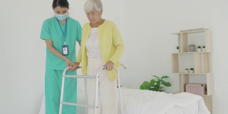 微笑的亚洲护士协助老年妇女起床。照顾护士支持病人从床上起来，并在家中走向助行器。帮助残疾老妇人站起来。