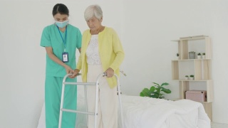 微笑的亚洲护士协助老年妇女起床。照顾护士支持病人从床上起来，并在家中走向助行器。帮助残疾老妇人站起来。视频素材模板下载