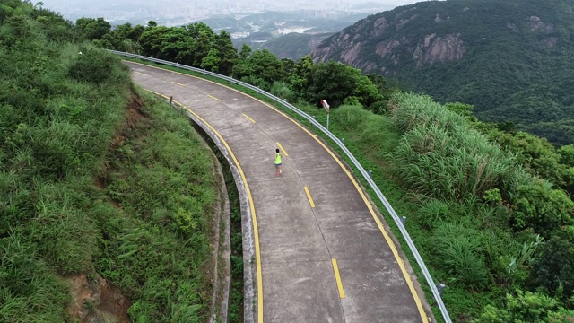 航拍健身女跑步者在山顶步道上奔跑，在山脚下远处的现代城市，慢镜头