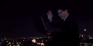 4K年轻的亚洲商人使用笔记本电脑与互联网视频会议与商业团队头脑风暴的想法，在城市的夜晚灯光背景下的办公楼成功的项目