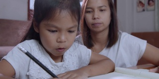 年轻的亚洲母亲在家里帮助她的女儿做家庭作业的慢动作。居家教育理念。