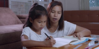 年轻的亚洲母亲在家里帮助她的女儿做家庭作业的慢动作。居家教育理念。