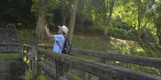 亚洲女游客戴着草帽和背包走在旧混凝土桥上，用手机拍照。游客喜欢在夏日的阳光下在森林公园里度过时光。