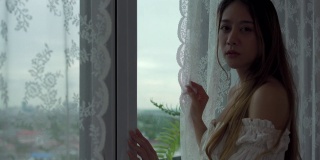 4K沮丧年轻美丽的亚洲女人独自住在公寓的窗户旁。孤独悲伤的女孩站在一边，看着窗外的城市，思考着生活中的问题。负面情绪的概念。