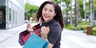 微笑年轻的亚洲女人与购物彩袋在商场背景。用智能手机在网上购物，站在商场大楼里微笑。生活方式的概念。慢镜头4 k。