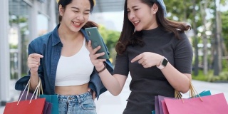 微笑年轻的亚洲女人与购物彩袋在商场背景。用智能手机在网上购物，站在商场大楼里微笑。生活方式的概念。慢镜头4 k。