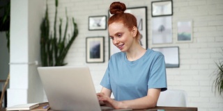 一个年轻的红发女人面带微笑，在家庭办公室的桌子上用笔记本电脑打字。