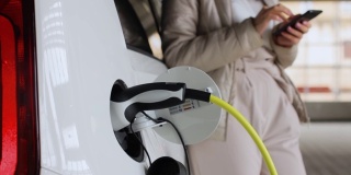 女人用手机附近充电的电动汽车;在户外公共充电站充电的车辆。汽车共享的概念
