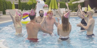 一群欢快的年轻人在水池中嬉戏。快乐的红发白人男子庆祝生日与多种族的朋友在夏季度假酒店。休闲的生活方式。