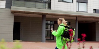 追踪慢动作拍摄的十几岁女食品快递员穿着绿色制服，带着保暖袋走在街上用智能手机上的导航应用程序，按门铃进入大楼