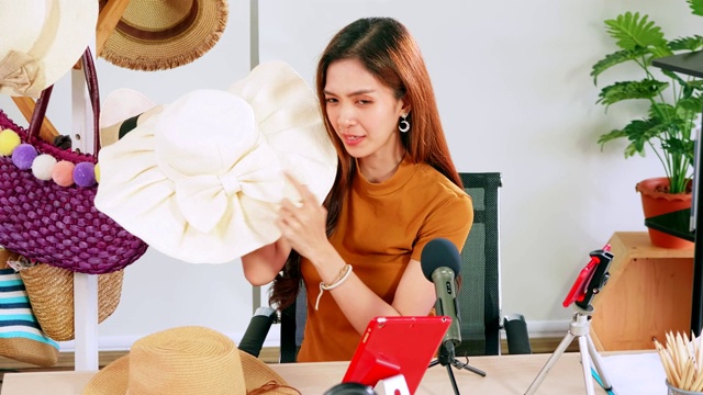年轻的亚洲女孩在社交媒体上通过在家直播销售时尚帽子和包。