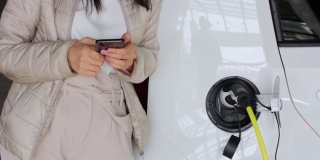 一名女子拿着智能手机在附近为电动车充电并检查电池状态。在户外公共充电站充电的车辆。汽车共享的概念