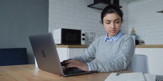 一位年轻的印度妇女在家工作，在电脑上做事，在笔记本上写笔记。她穿着一件衬衫和一件毛衣