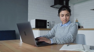 一位年轻的印度妇女在家工作，在电脑上做事，在笔记本上写笔记。她穿着一件衬衫和一件毛衣视频素材模板下载