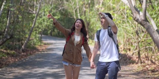 快乐的亚洲年轻情侣背着背包，在暑假里一起走在森林土路上的高清慢镜头。面带微笑的男女朋友喜欢在自然小道上徒步旅行。