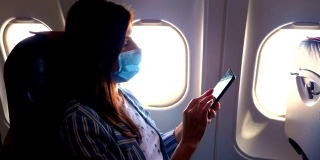 一名年轻女子戴着防护面具，坐在飞机内靠近照明装置的地方使用手机和智能手机。新冠肺炎疫情后恢复航班。开放边界。Post-quarantine旅行