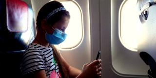 女孩、孩子戴着防护面具在飞机内使用手机、智能手机，坐在照明装置附近。新冠肺炎疫情后恢复航班。开放边界。Post-quarantine旅行