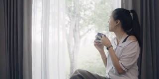 亚洲幸福的女人使用智能手机和喝咖啡在客厅的沙发在家里的背景。以快乐放松为休闲活动理念。4 k慢动作。