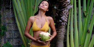 美丽的年轻亚洲女人在黄色比基尼在热带度假花园喝新鲜的年轻椰子。暑假