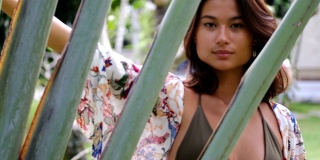 美丽的年轻亚洲女人穿着比基尼和夏天的海滩装在绿色的棕榈树后面的热带花园。暑假