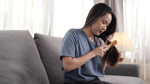 年轻的亚洲女子坐在家里客厅的沙发上梳头，是人们的生活方式发型师的概念。