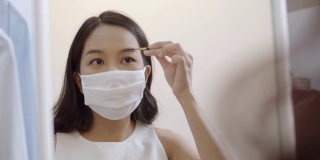 年轻的亚洲妇女戴着防护口罩，在隔离期间画眉毛。