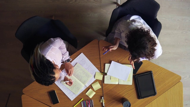 俯视图女性的手写笔记在记事本。写下商业想法。两个商业女人讨论想法的特写。穿着白衬衫的妇女坐在木桌旁，面目全非