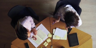 俯视图女性的手写笔记在记事本。写下商业想法。两个商业女人讨论想法的特写。穿着白衬衫的妇女坐在木桌旁，面目全非