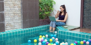 一个穿着休闲服在笔记本电脑上工作的女人，早上坐在游泳池边喝咖啡，把腿放在水里放松在一个豪华度假酒店。概念商务人士在周末活动。