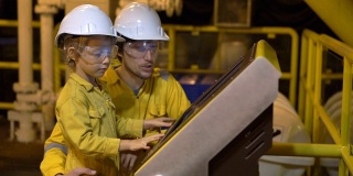 一个年轻的男子和一个小男孩都穿着黄色的工作服、眼镜和头盔在一个工业环境、石油平台或液化气工厂看着屏幕。Slowmotion拍摄