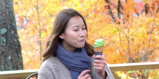 微笑的亚洲年轻女性游客在公园里吃日本抹茶绿茶冰淇淋的4K肖像。快乐的女士，放松和享受清新寒冷的天气，美丽的秋天在日本。